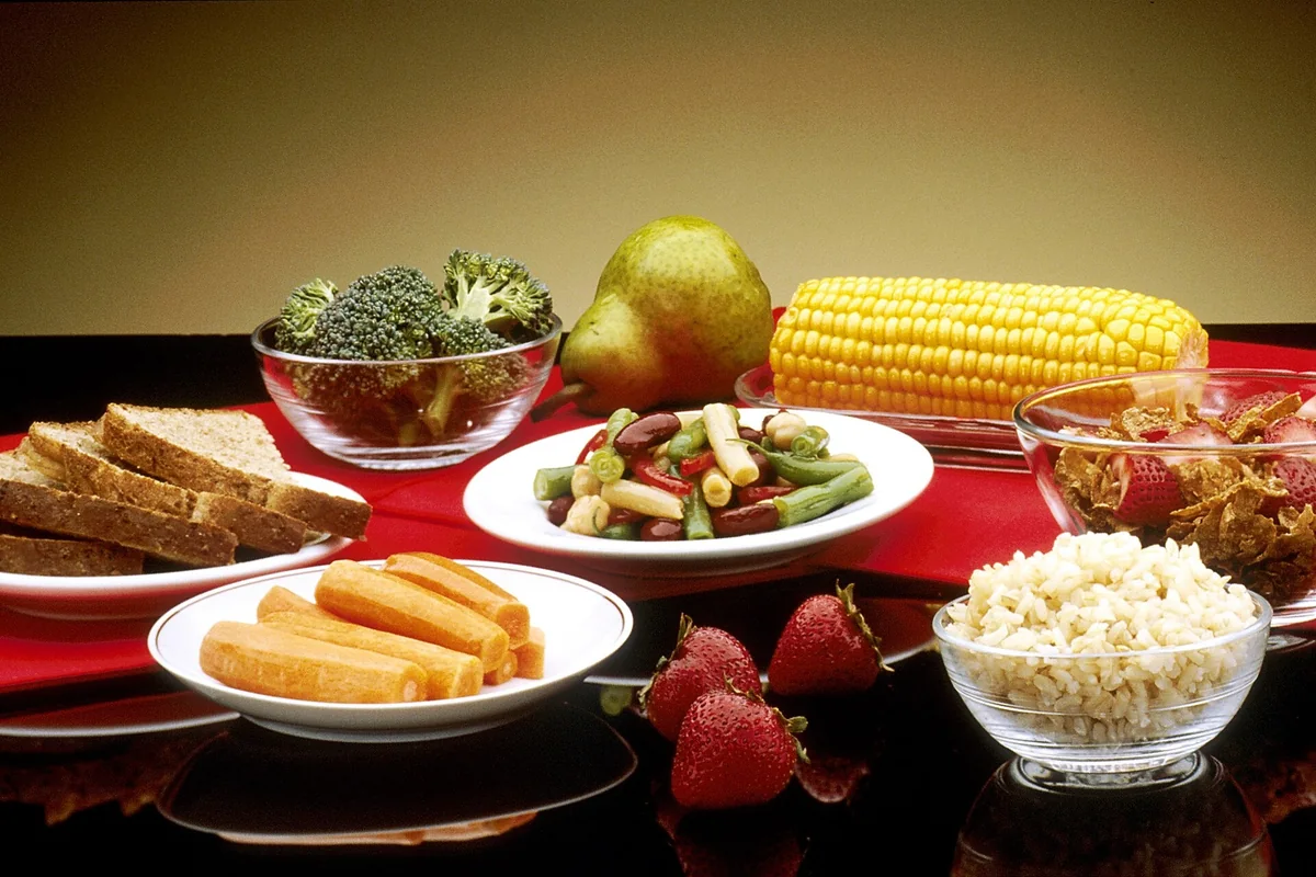 Farfurii și boluri cu broccoli, pară, porumb, pâine integrală, salată de fasole, orez și căpșuni, pe o față de masă roșie