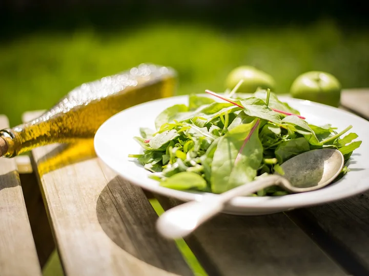 Farfurie albă cu salată verde, pe o masă de lemn, lângă o sticlă cu ulei și două mere verzi