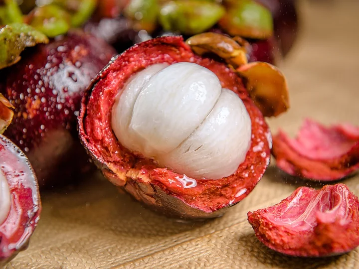 Fruct de mangostan deschis, cu pulpa albă, în segmente, într-o coajă roșie, lângă bucăți de coajă