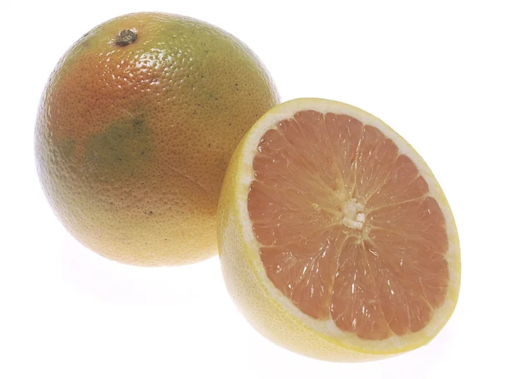 Un grapefruit întreg, cu coaja galbenă, cu nuanțe de portocaliu și verde, lângă un grapefruit tăiat în două, cu pulpa roz-pal, în segmente