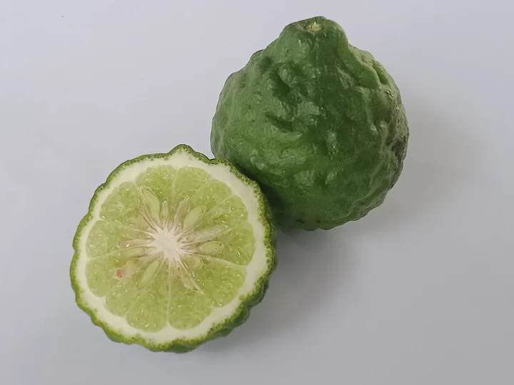 Fruct de lime kaffir, cu coaja verde-închis și cocoloașe, lângă o jumătate tăiată, cu miez verzui