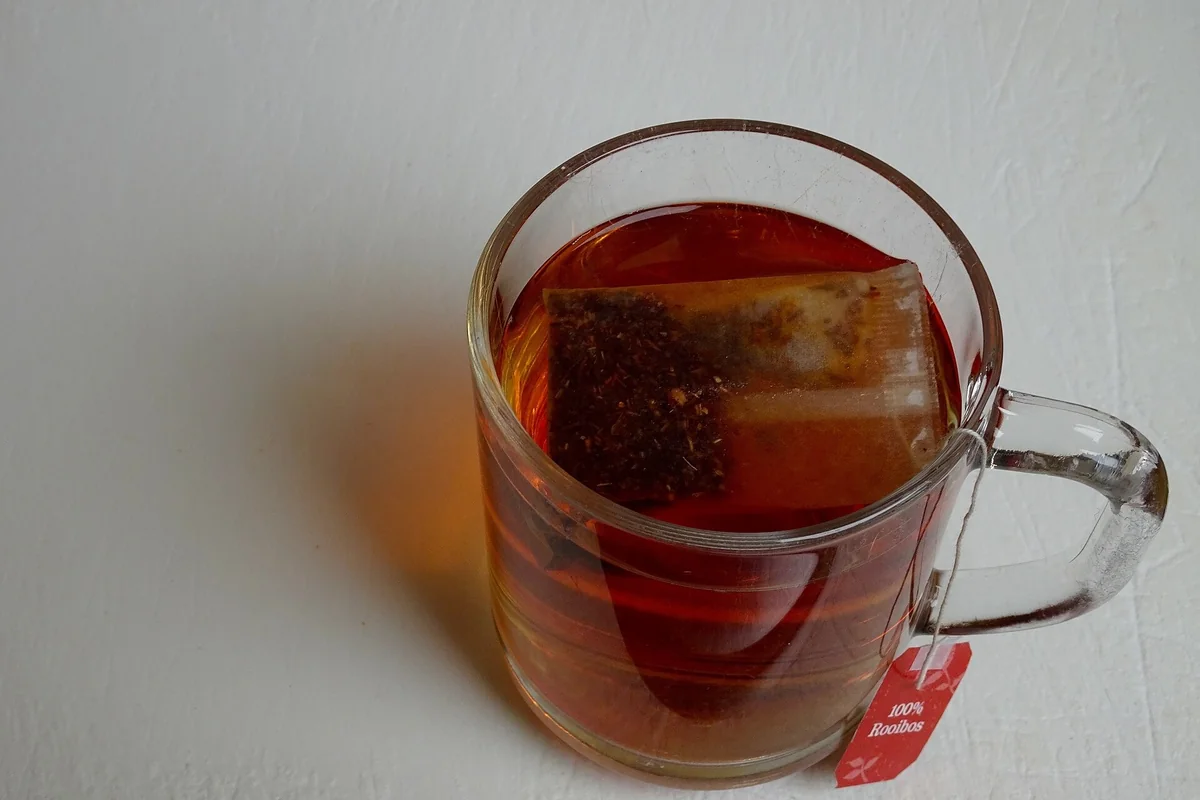 Cană de sticlă cu ceai roșu-brun de rooibos, cu un plic de ceai scufundat și cu eticheta roșie „100% Rooibos” atârnată lângă cană, pe fond alb